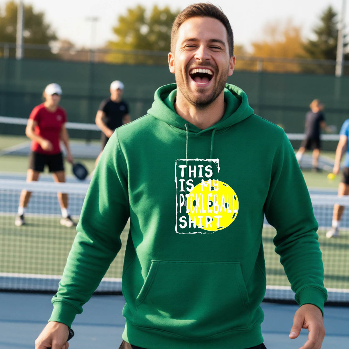 A man wearing a green hoodie with a yellow and white graphic that reads 'THIS IS MY PICKLEBALL SHIRT' stands on a pickleball court. 