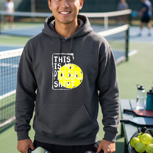 A man wearing a dark gray hoodie with a yellow and white graphic that reads 'THIS IS MY PICKLEBALL SHIRT' stands on a pickleball court. 
