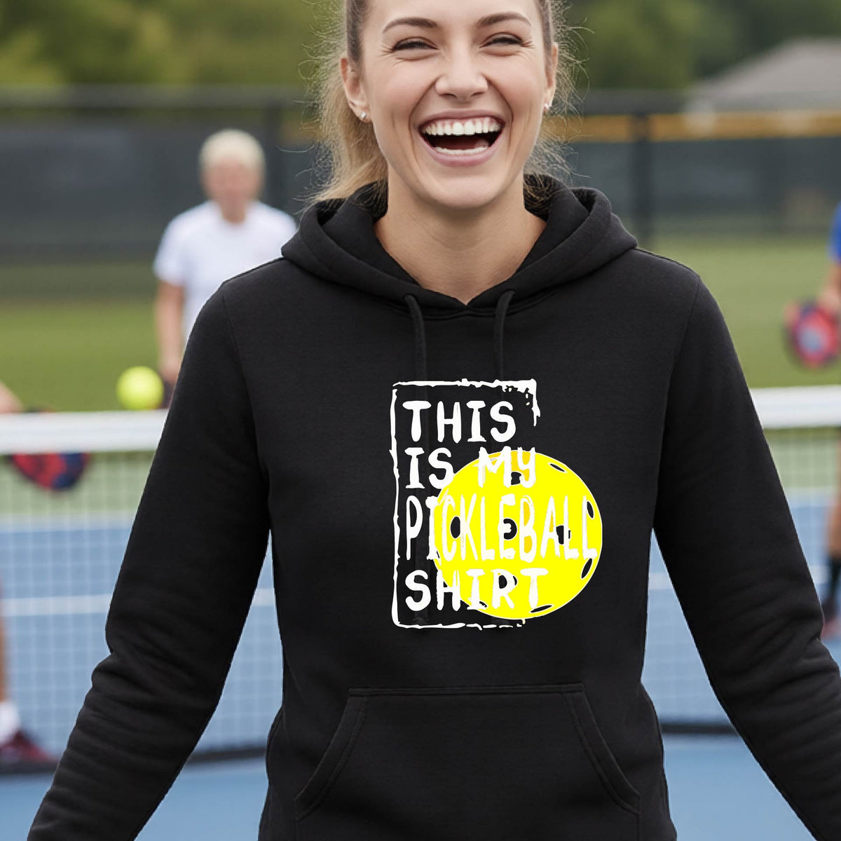 A smiling blonde woman wearing a black pullover hoodie with a graphic print that reads 'THIS IS MY PICKLEBALL SHIRT' in white text over a bright yellow pickleball, standing on a pickleball court.
