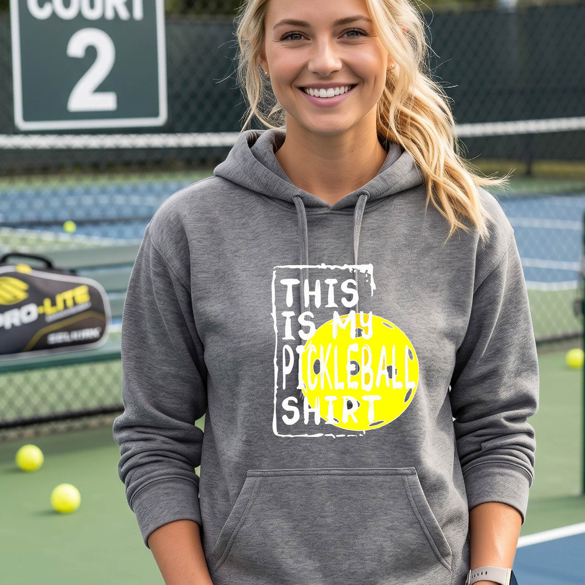 A smiling blonde woman wearing a heather gray pullover hoodie with a graphic print that reads 'THIS IS MY PICKLEBALL SHIRT' in white text over a bright yellow pickleball, standing on a pickleball court.