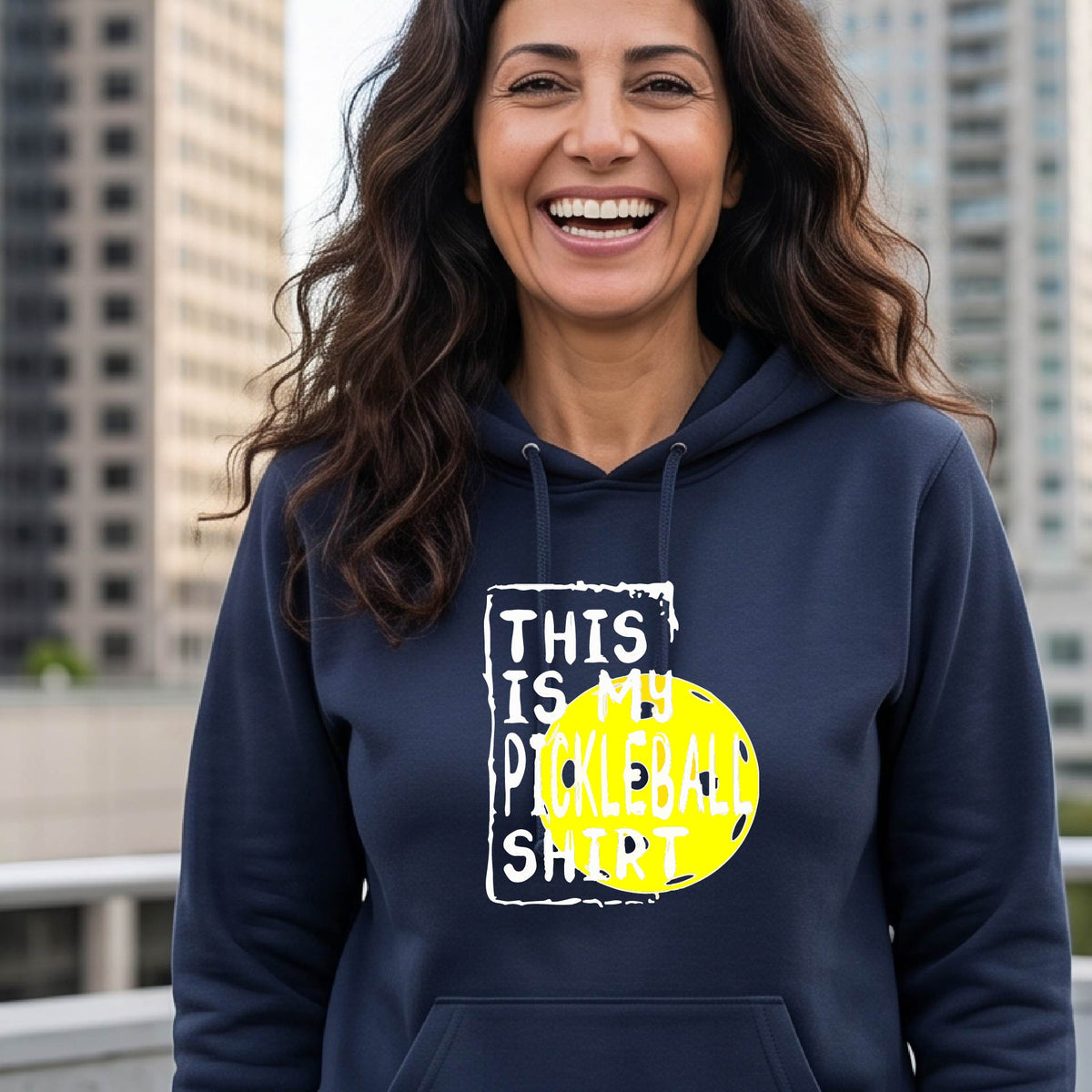A smiling blonde woman wearing a navy blue pullover hoodie with a graphic print that reads 'THIS IS MY PICKLEBALL SHIRT' in white text over a bright yellow pickleball, standing on a pickleball court.