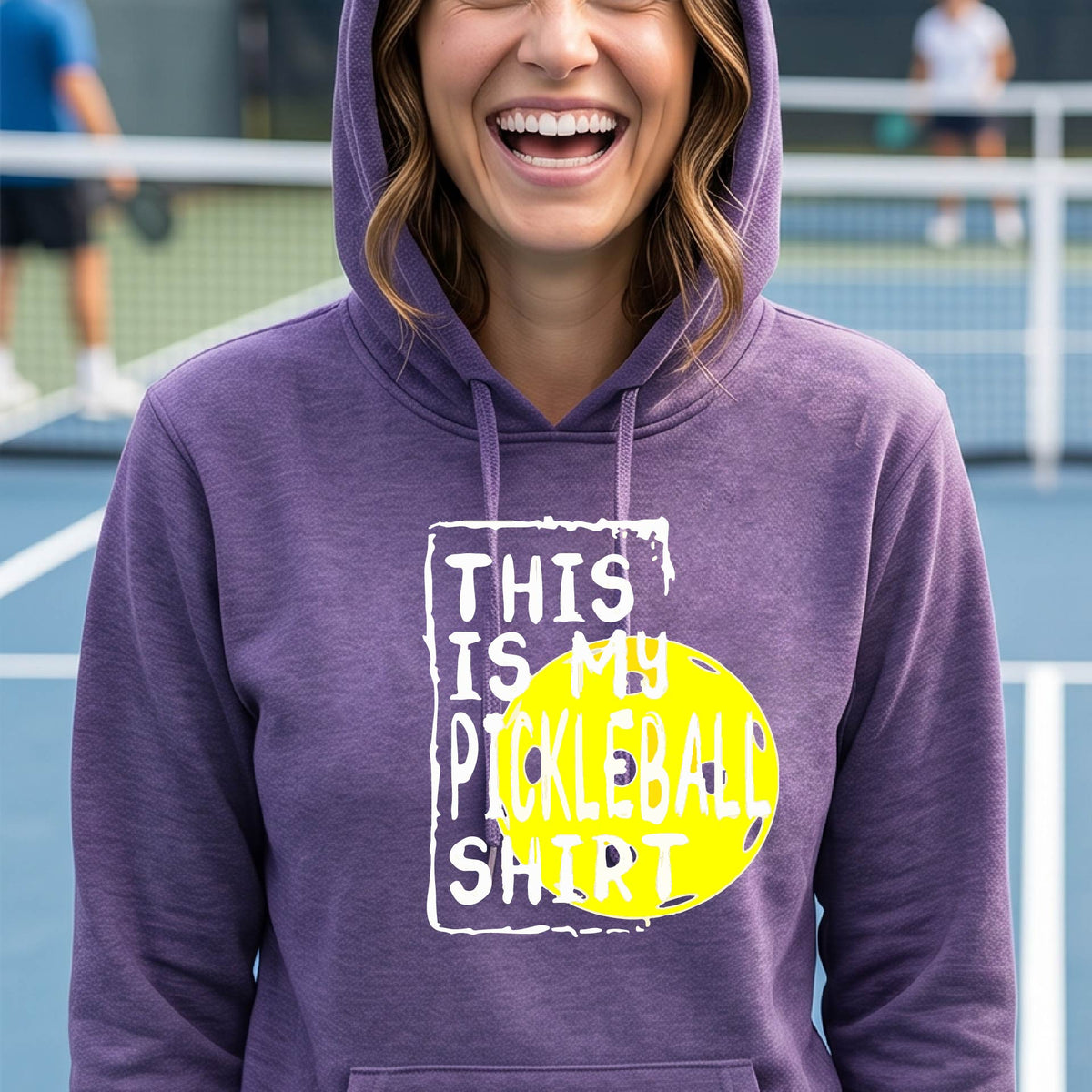 A smiling blonde woman wearing a purple pullover hoodie with a graphic print that reads 'THIS IS MY PICKLEBALL SHIRT' in white text over a bright yellow pickleball, standing on a pickleball court.