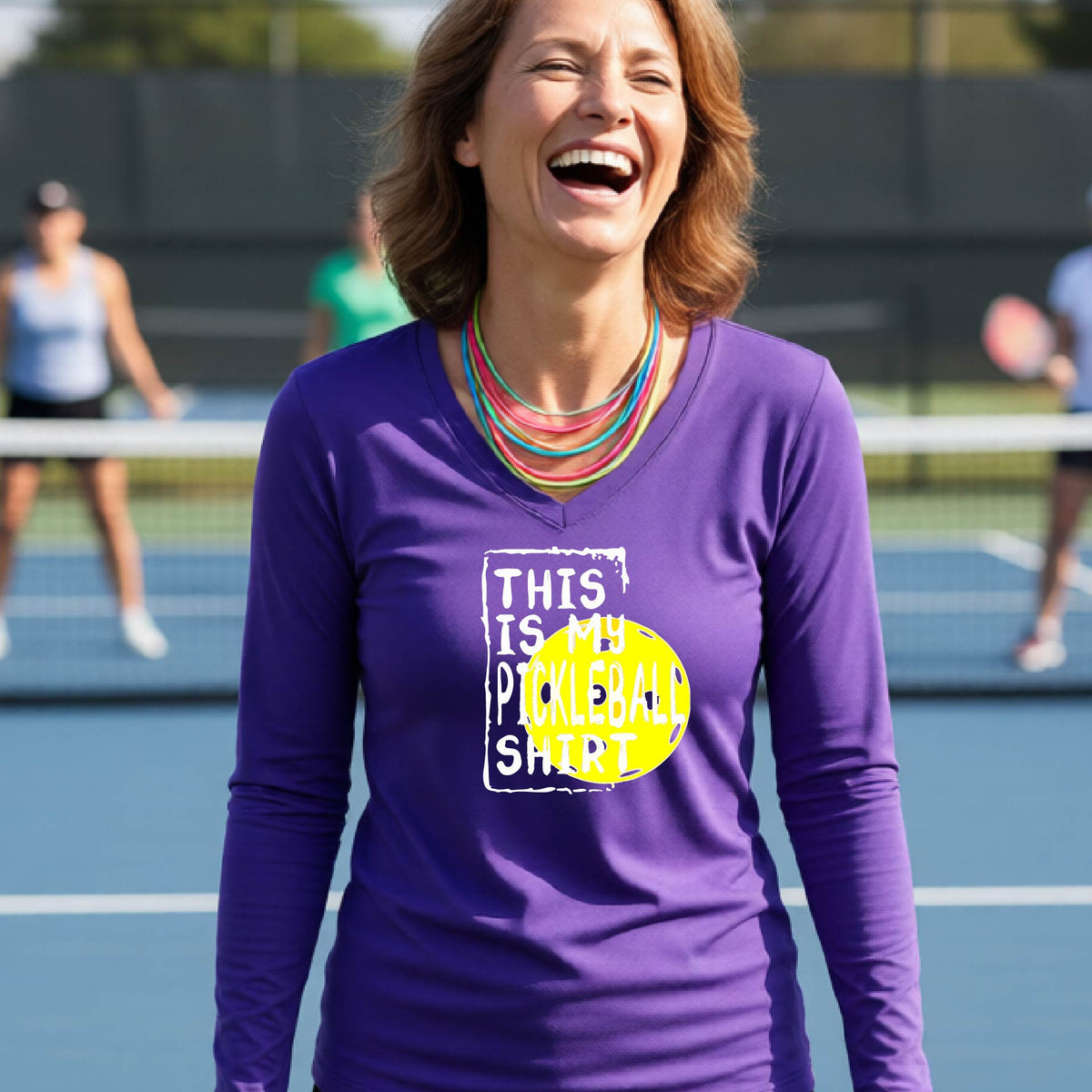 A woman laughing on an outdoor pickleball court wearing a purple long-sleeve t-shirt with a graphic that reads 'THIS IS MY PICKLEBALL SHIRT' in white and a yellow pickleball graphic.