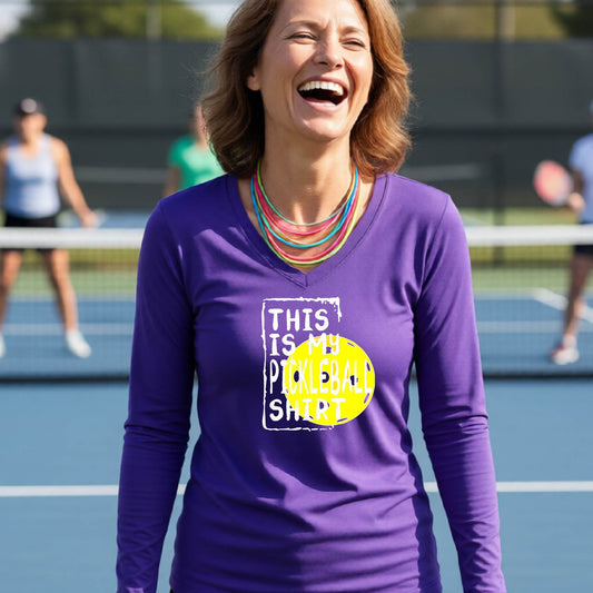 A woman laughing on an outdoor pickleball court wearing a purple long-sleeve t-shirt with a graphic that reads 'THIS IS MY PICKLEBALL SHIRT' in white and a yellow pickleball graphic.