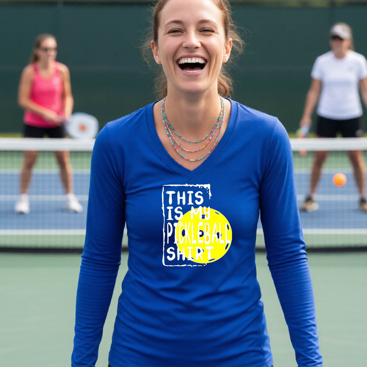 A woman laughing on an outdoor pickleball court wearing a royal blue long-sleeve t-shirt with a graphic that reads 'THIS IS MY PICKLEBALL SHIRT' in white and a yellow pickleball graphic.