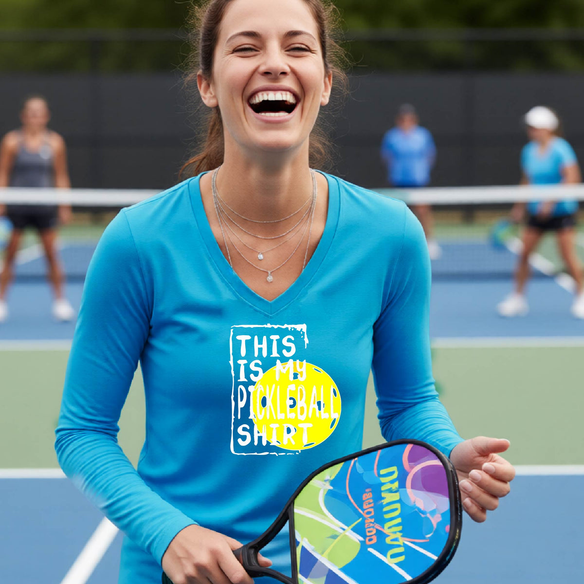 A woman laughing on an outdoor pickleball court wearing a bright blue long-sleeve t-shirt with a graphic that reads 'THIS IS MY PICKLEBALL SHIRT' in white and a yellow pickleball graphic.