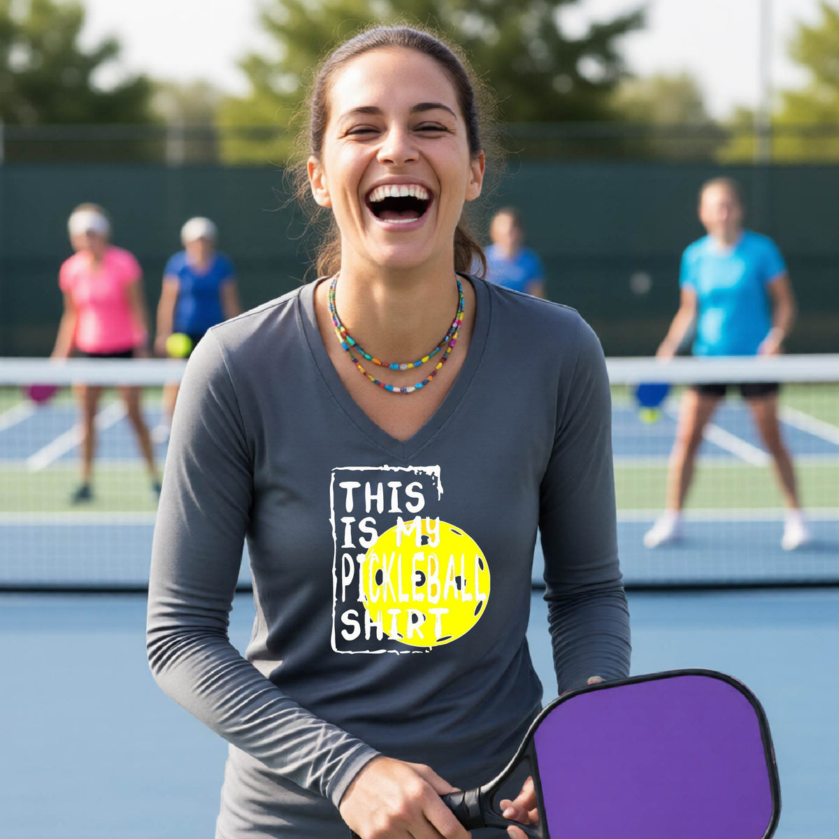A woman laughing on an outdoor pickleball court wearing a gray long-sleeve t-shirt with a graphic that reads 'THIS IS MY PICKLEBALL SHIRT' in white and a yellow pickleball graphic.