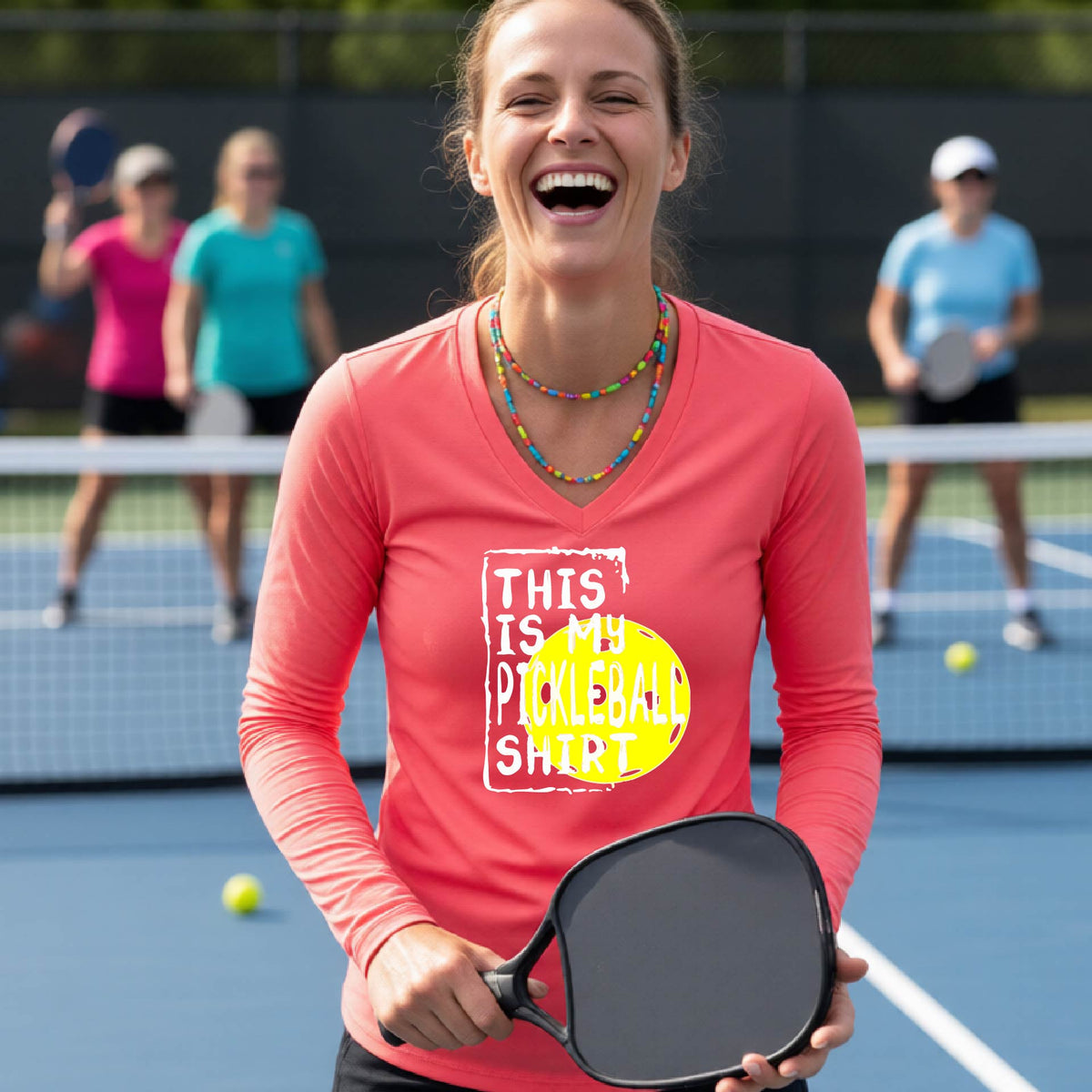 A woman laughing on an outdoor pickleball court wearing a coral pink long-sleeve t-shirt with a graphic that reads 'THIS IS MY PICKLEBALL SHIRT' in white and a yellow pickleball graphic.
