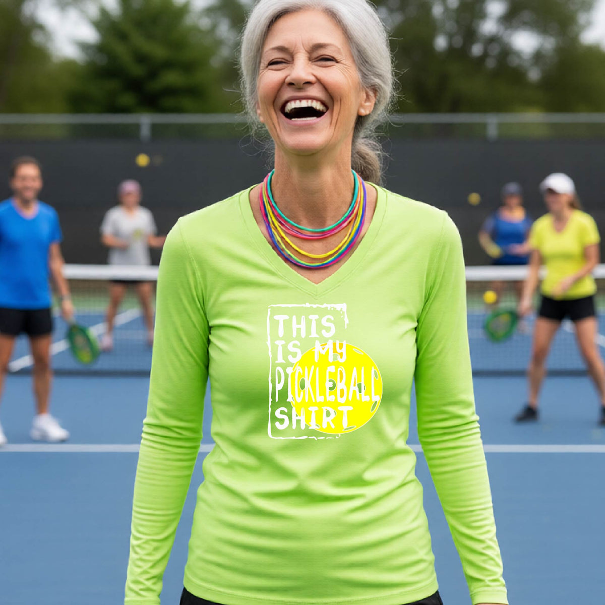 A woman laughing on an outdoor pickleball court wearing a lime green long-sleeve t-shirt with a graphic that reads 'THIS IS MY PICKLEBALL SHIRT' in white and a yellow pickleball graphic.