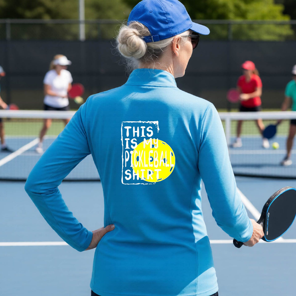 A woman facing backwards on an outdoor pickleball court wearing a bright blue quarter-zip pullover shirt with a graphic that reads 'THIS IS MY PICKLEBALL SHIRT' in white with a yellow pickleball graphic on the back.