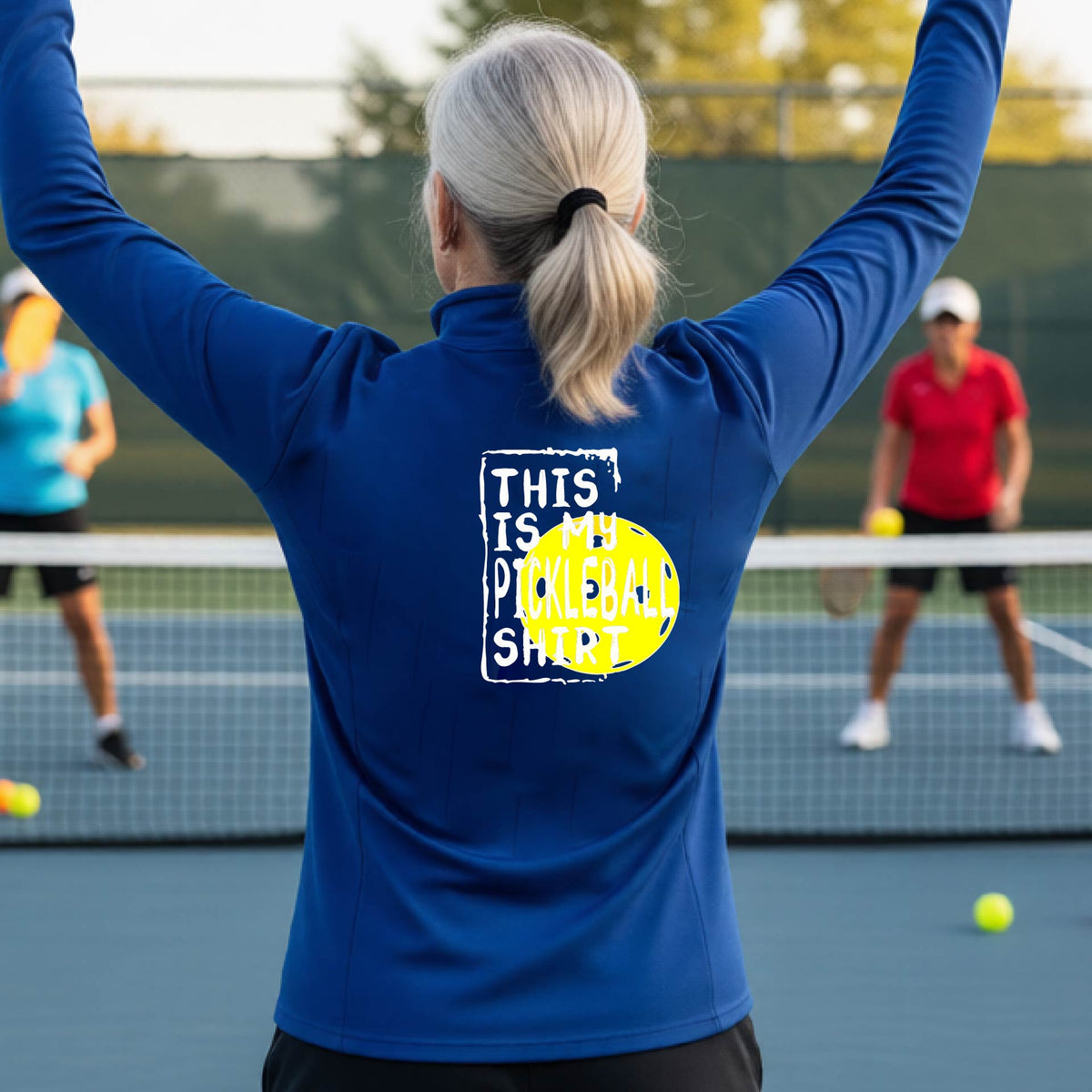 A woman facing backwards on an outdoor pickleball court wearing a royal blue quarter-zip pullover shirt with a graphic that reads 'THIS IS MY PICKLEBALL SHIRT' in white with a yellow pickleball graphic on the back.