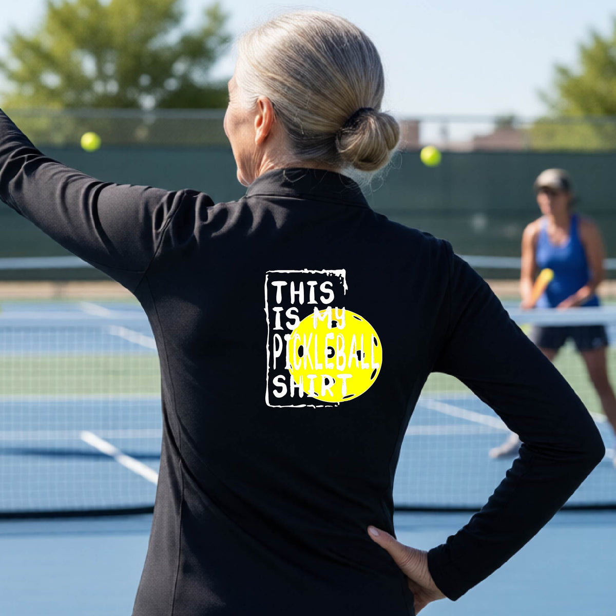 A woman facing backwards on an outdoor pickleball court wearing a black quarter-zip pullover shirt with a graphic that reads 'THIS IS MY PICKLEBALL SHIRT' in white with a yellow pickleball graphic on the back.