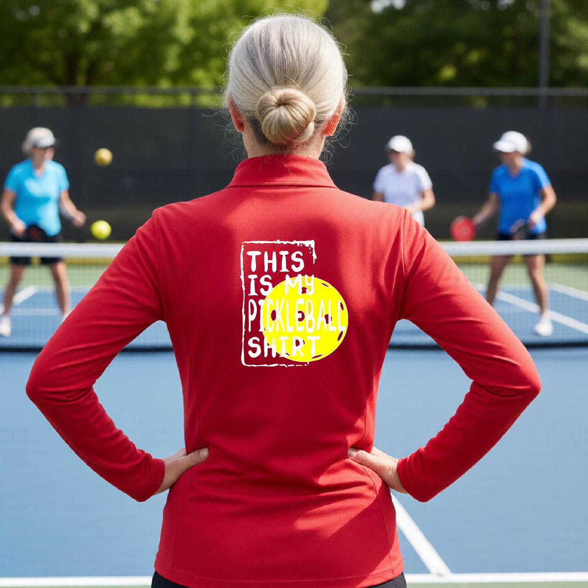 A woman facing backwards on an outdoor pickleball court wearing a red quarter-zip pullover shirt with a graphic that reads 'THIS IS MY PICKLEBALL SHIRT' in white with a yellow pickleball graphic on the back.