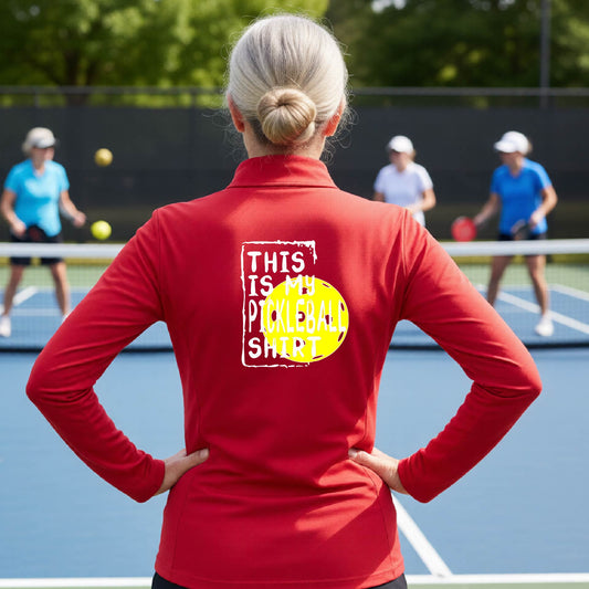 A woman facing backwards on an outdoor pickleball court wearing a red quarter-zip pullover shirt with a graphic that reads 'THIS IS MY PICKLEBALL SHIRT' in white with a yellow pickleball graphic on the back.