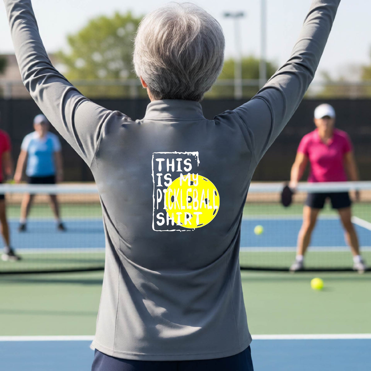 A woman facing backwards on an outdoor pickleball court wearing a gray quarter-zip pullover shirt with a graphic that reads 'THIS IS MY PICKLEBALL SHIRT' in white with a yellow pickleball graphic on the back.