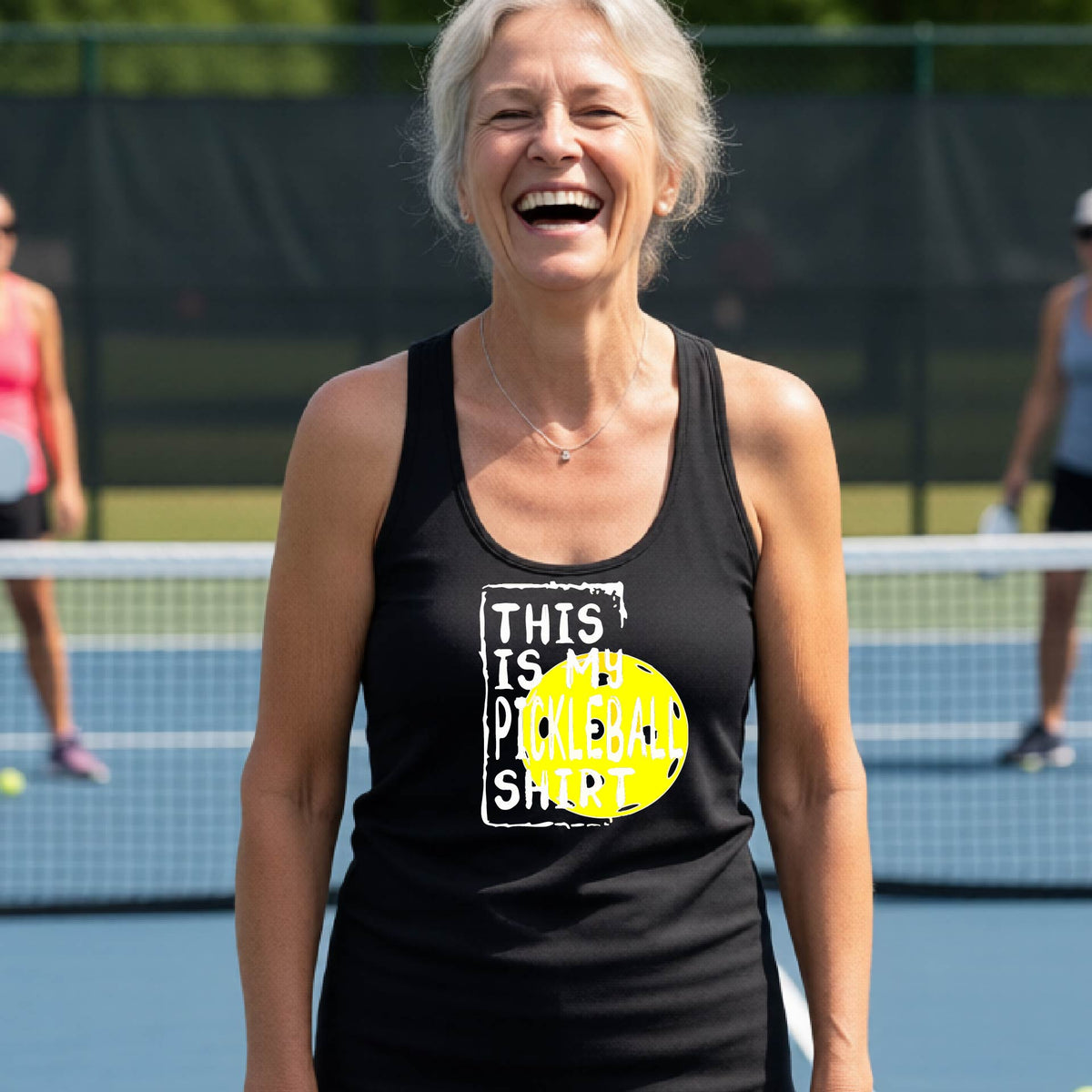 A woman laughing on a pickleball court wearing a black tank top with a graphic that reads 'THIS IS MY PICKLEBALL SHIRT' in white with a yellow pickelball graphic.