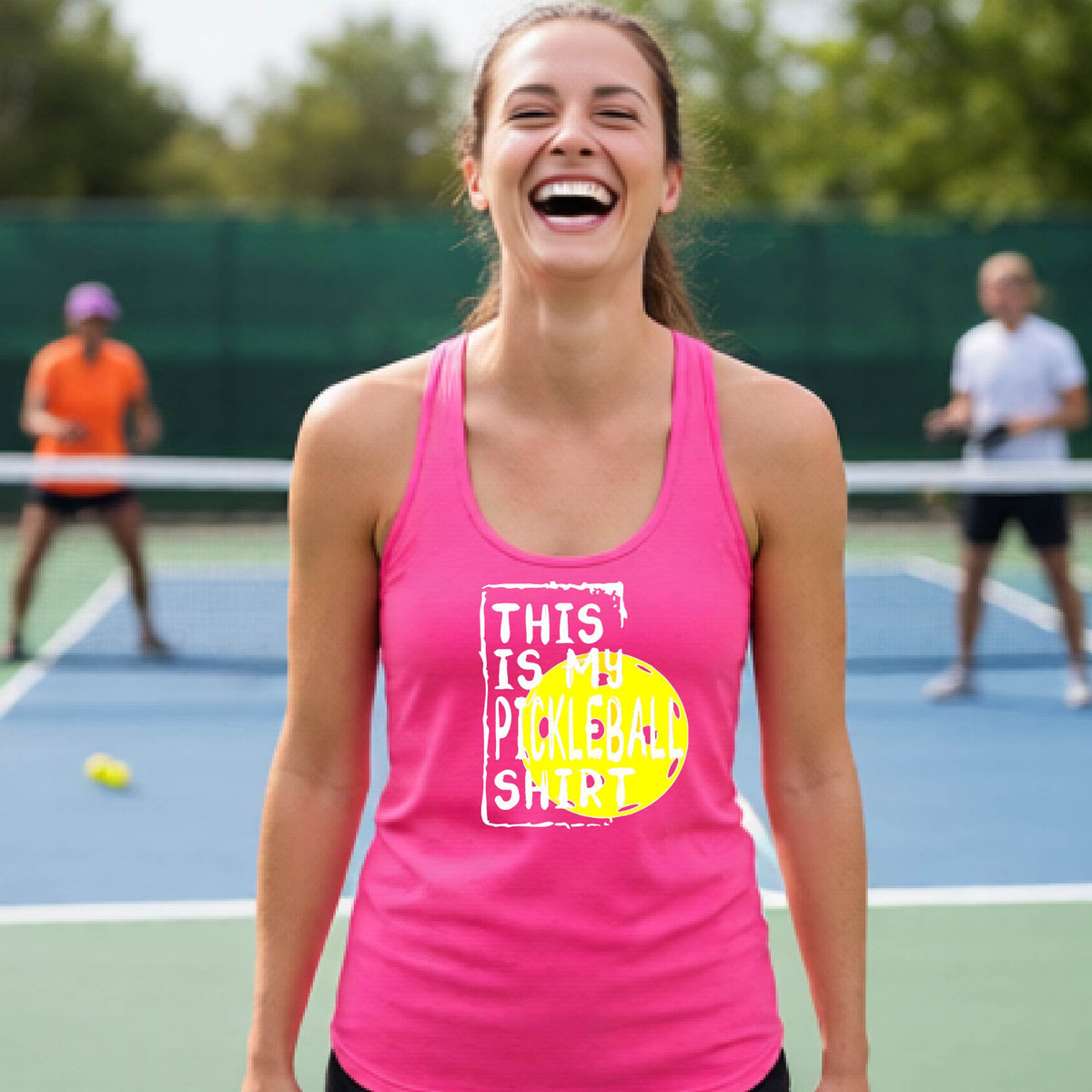 A woman laughing on a pickleball court wearing a pink tank top with a graphic that reads 'THIS IS MY PICKLEBALL SHIRT' in white with a yellow pickelball graphic.