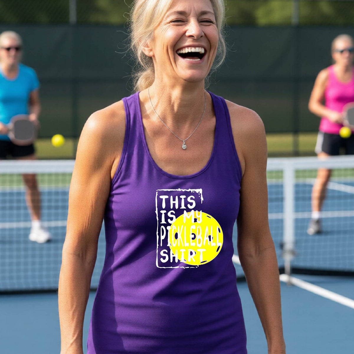 A woman laughing on a pickleball court wearing a purple tank top with a graphic that reads 'THIS IS MY PICKLEBALL SHIRT' in white with a yellow pickelball graphic.
