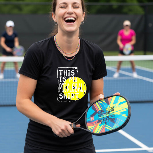 A woman laughing on a pickleball court wearing a black graphic t-shirt that reads 'THIS IS MY PICKLEBALL SHIRT' over a yellow pickleball graphic.