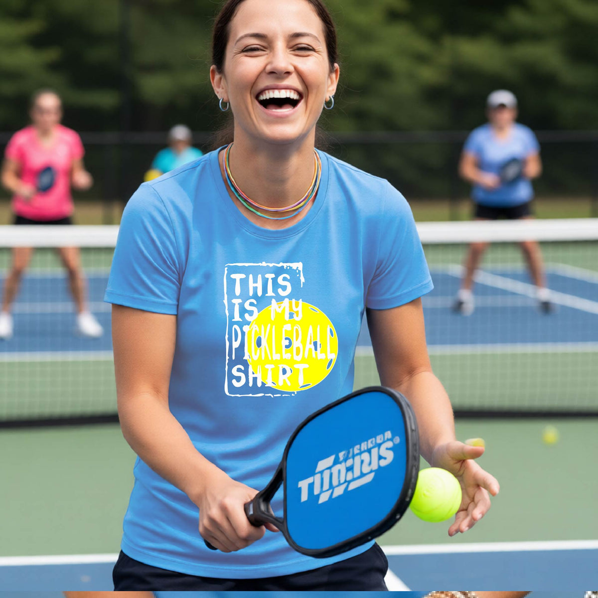 A woman laughing on a pickleball court wearing a blue graphic t-shirt that reads 'THIS IS MY PICKLEBALL SHIRT' over a yellow pickleball graphic.