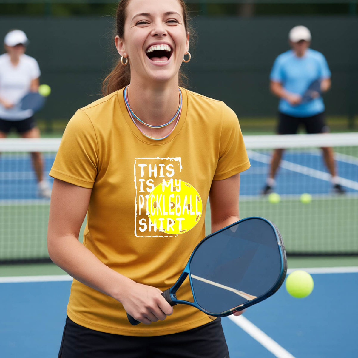 A woman laughing on a pickleball court wearing a gold graphic t-shirt that reads 'THIS IS MY PICKLEBALL SHIRT' over a yellow pickleball graphic.