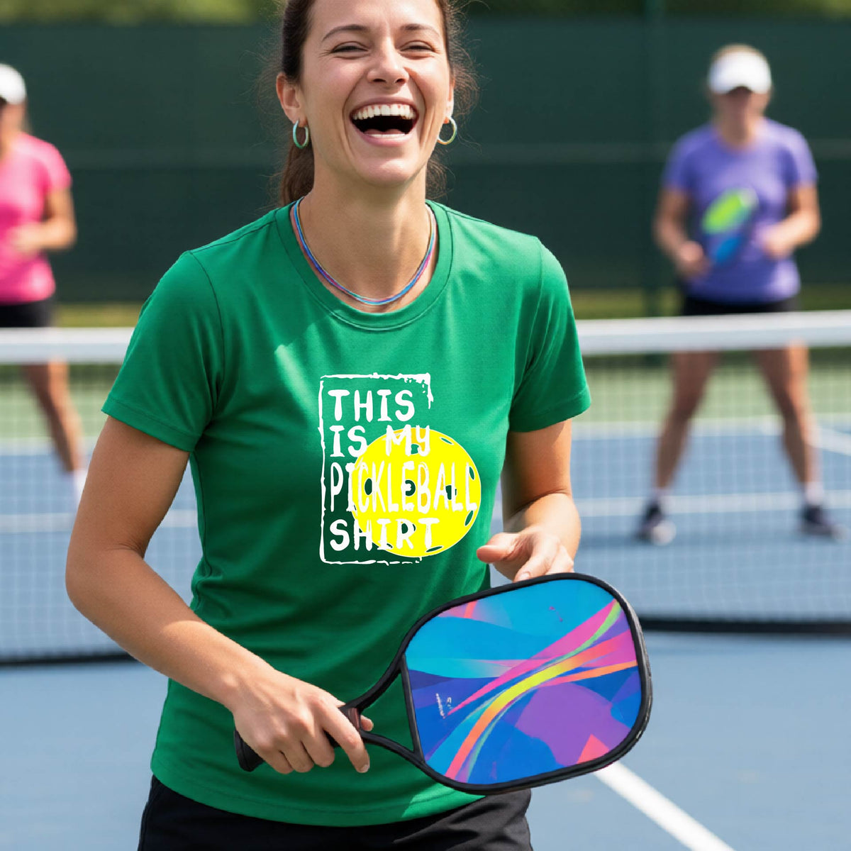 A woman laughing on a pickleball court wearing a green graphic t-shirt that reads 'THIS IS MY PICKLEBALL SHIRT' over a yellow pickleball graphic.