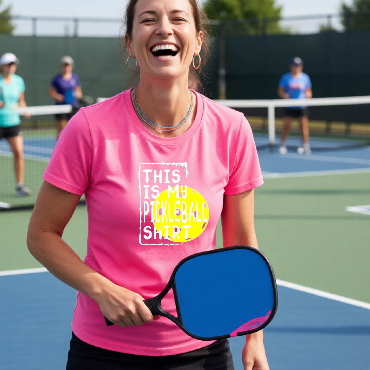 A woman laughing on a pickleball court wearing a pink graphic t-shirt that reads 'THIS IS MY PICKLEBALL SHIRT' over a yellow pickleball graphic.