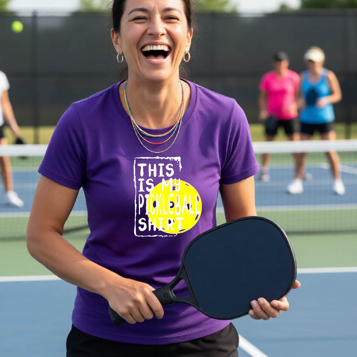 A woman laughing on a pickleball court wearing a purple graphic t-shirt that reads 'THIS IS MY PICKLEBALL SHIRT' over a yellow pickleball graphic.