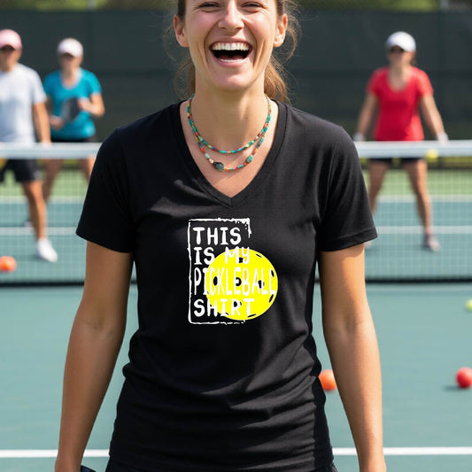 A woman on a pickleball court wearing a black V-neck t-shirt with a graphic that reads 'THIS IS MY PICKLEBALL SHIRT' in white text over a yellow pickleball.