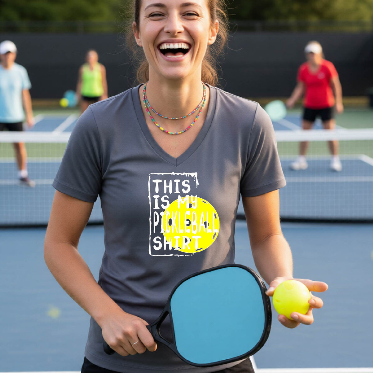 A woman on a pickleball court wearing a gray V-neck t-shirt with a graphic that reads 'THIS IS MY PICKLEBALL SHIRT' in white text over a yellow pickleball.