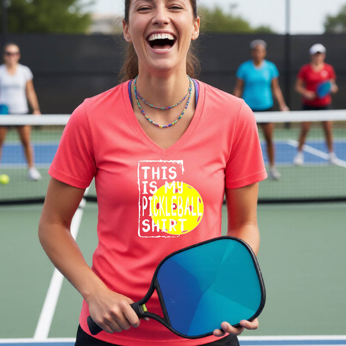 A woman on a pickleball court wearing a coral pink V-neck t-shirt with a graphic that reads 'THIS IS MY PICKLEBALL SHIRT' in white text over a yellow pickleball.