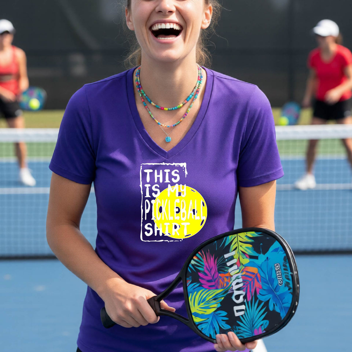 A woman on a pickleball court wearing a purple V-neck t-shirt with a graphic that reads 'THIS IS MY PICKLEBALL SHIRT' in white text over a yellow pickleball.