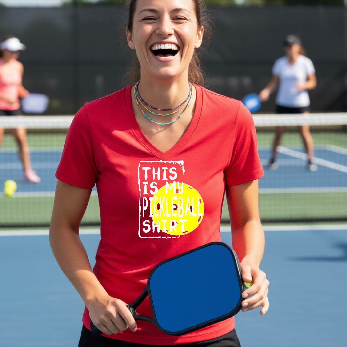 A woman on a pickleball court wearing a red V-neck t-shirt with a graphic that reads 'THIS IS MY PICKLEBALL SHIRT' in white text over a yellow pickleball.