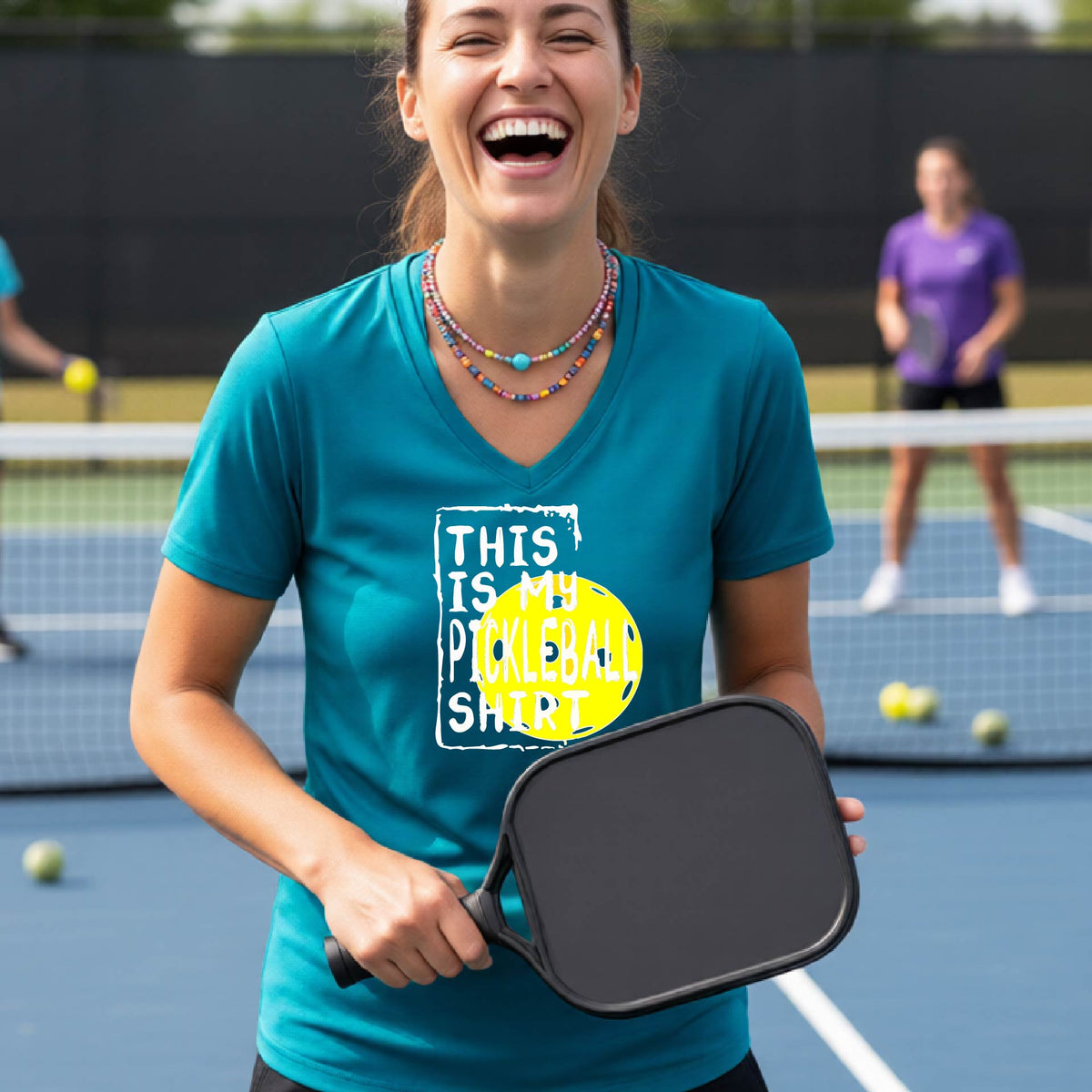 A woman on a pickleball court wearing a teal blue V-neck t-shirt with a graphic that reads 'THIS IS MY PICKLEBALL SHIRT' in white text over a yellow pickleball.