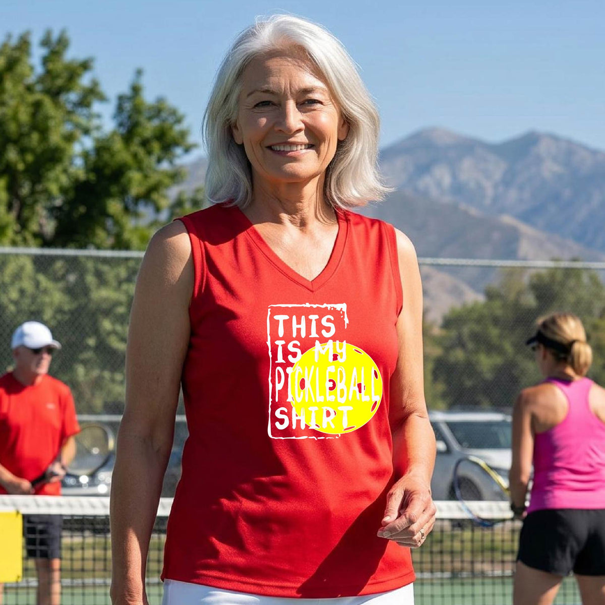 A woman on a pickleball court wearing a red tank top with a yellow and white graphic that reads 'THIS IS MY PICKLEBALL SHIRT' including the yellow pickleball icon.