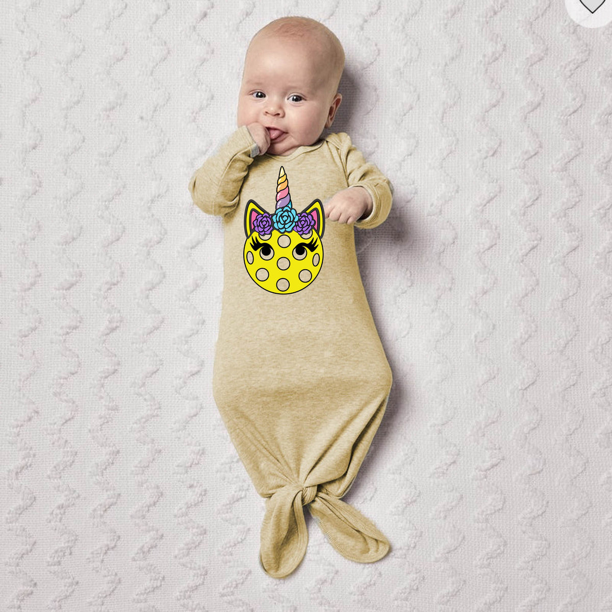A baby on a blanket wearing a beige bodysuit with a colorful pickleball unicorn graphic.