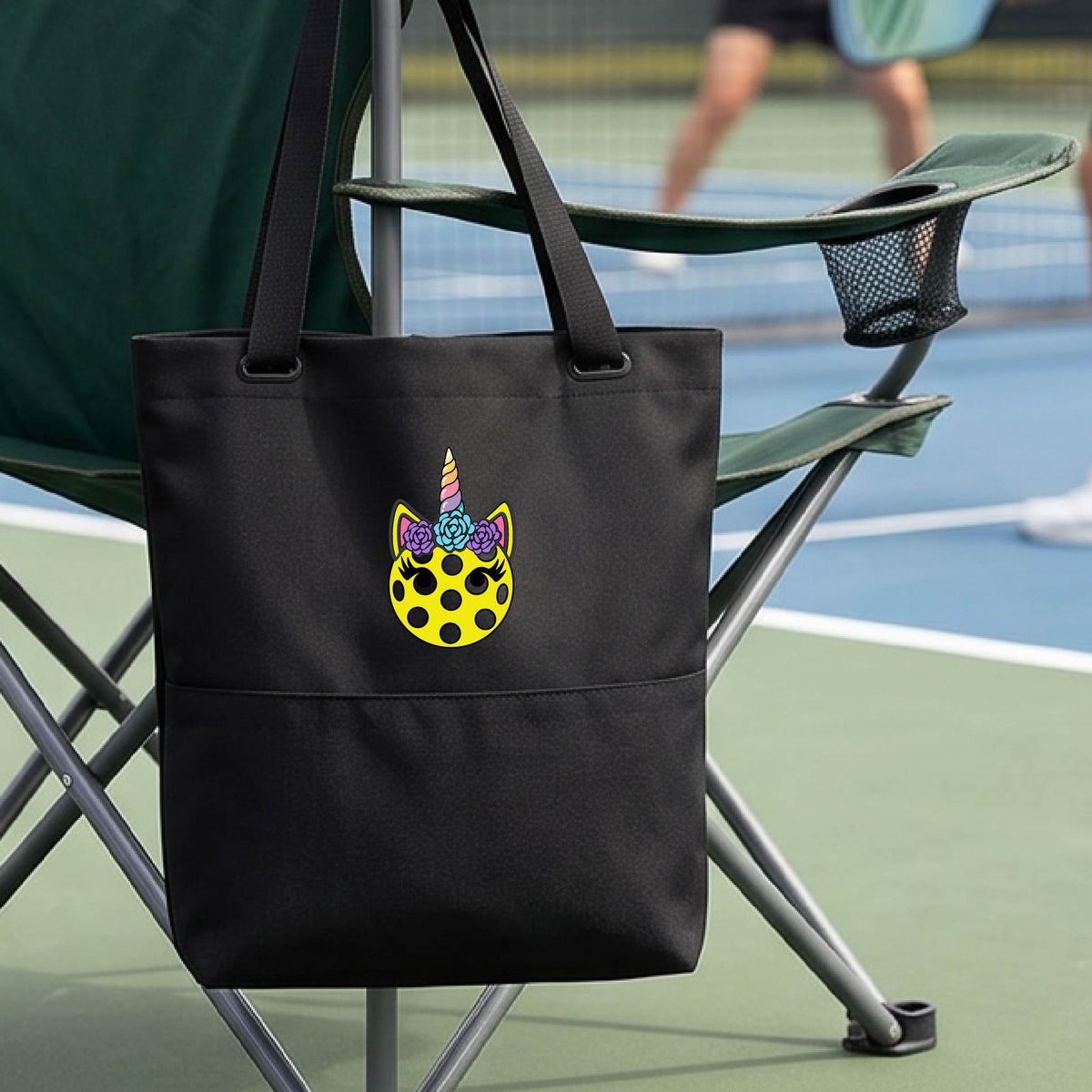 A close-up of a black medium size pickleball tote with a graphic of a yellow pickleball with unicorn ears and horns as a graphic on the front.