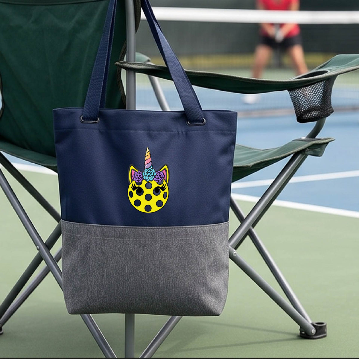 A close-up of a blue and gray medium size pickleball tote with a graphic of a yellow pickleball with unicorn ears and horns as a graphic on the front.
