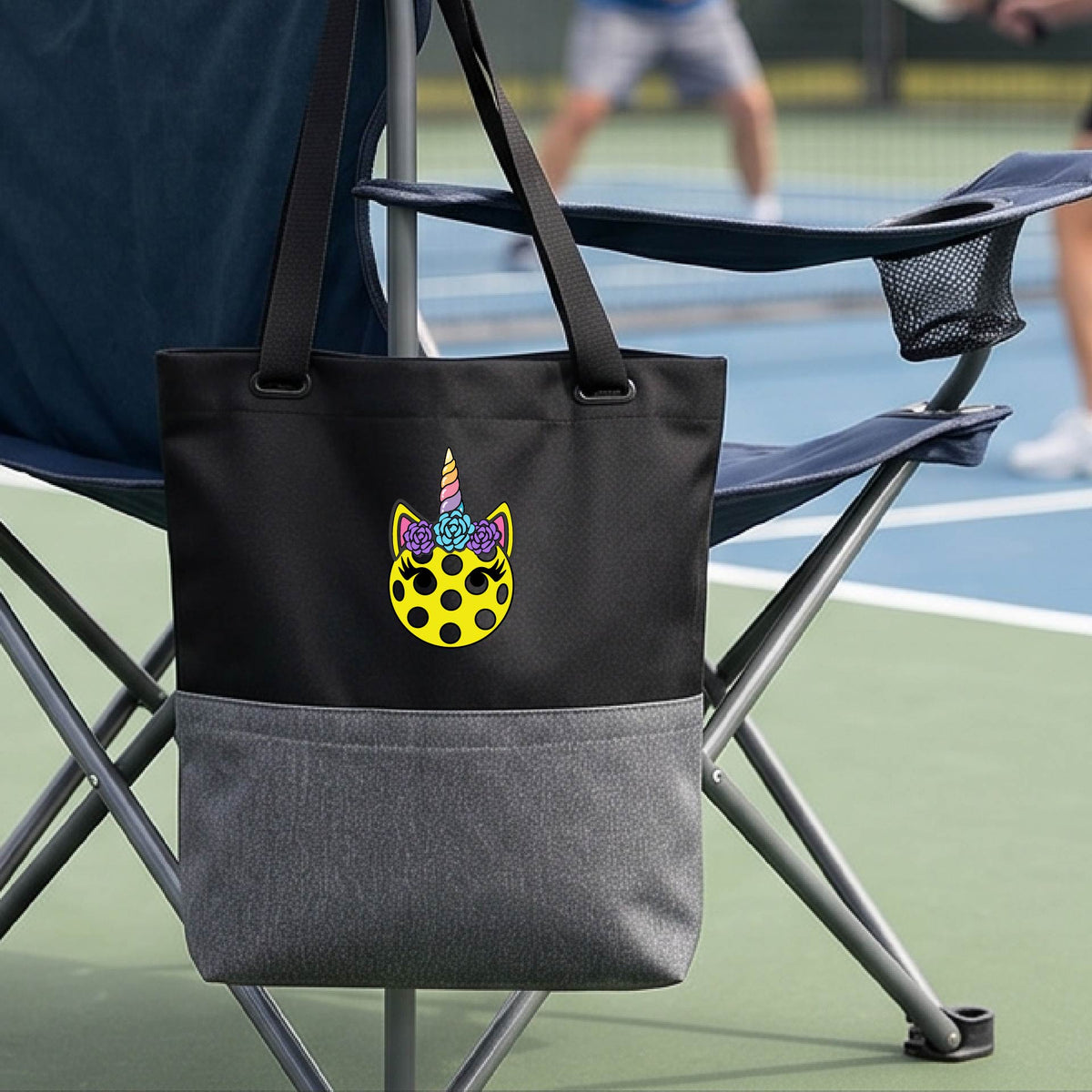 A close-up of a black and gray medium size pickleball tote with a graphic of a yellow pickleball with unicorn ears and horns as a graphic on the front.