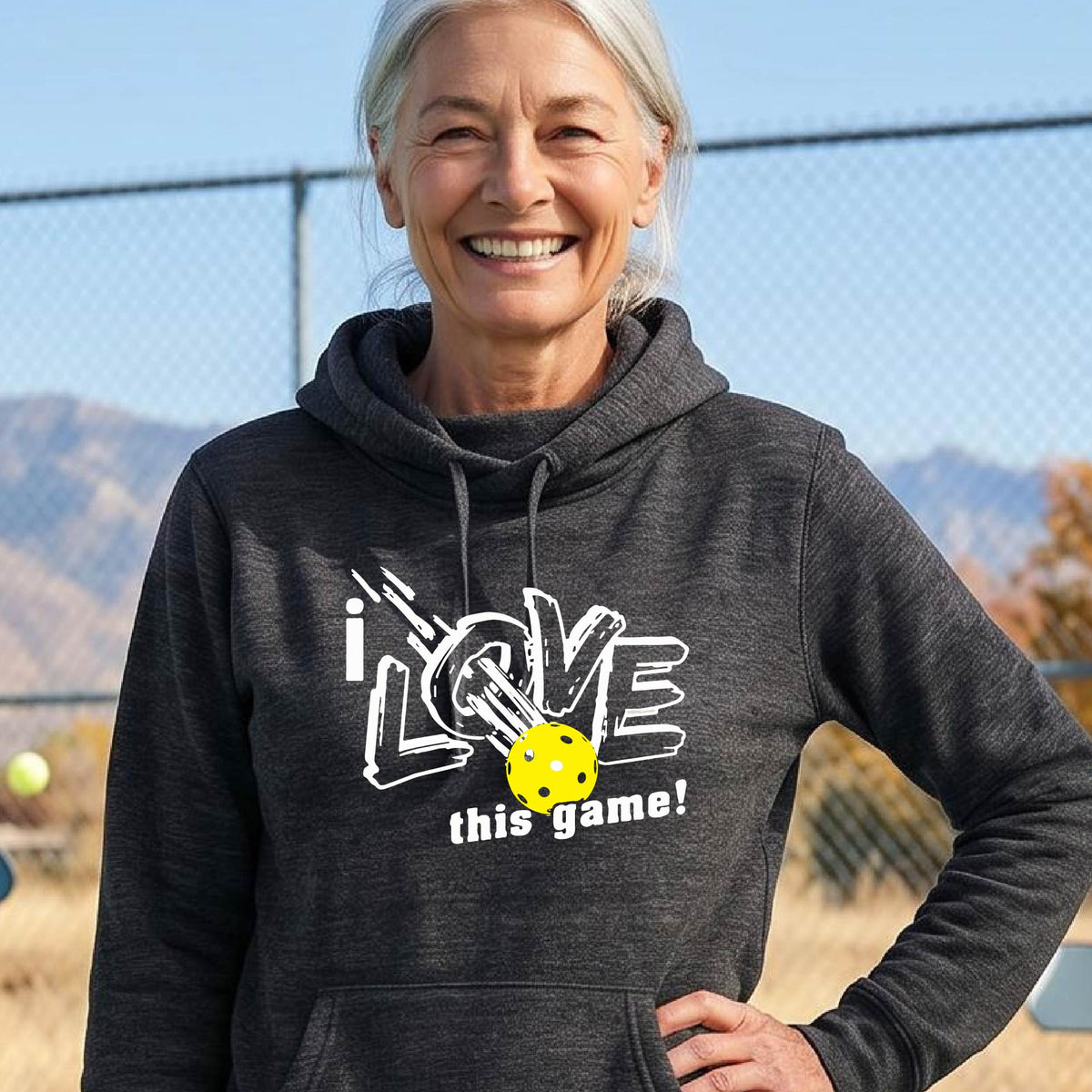 A woman wearing a black hoodie with a graphic that reads 'i LOVE this game!' in white, with a yellow pickleball over the letter O. 
