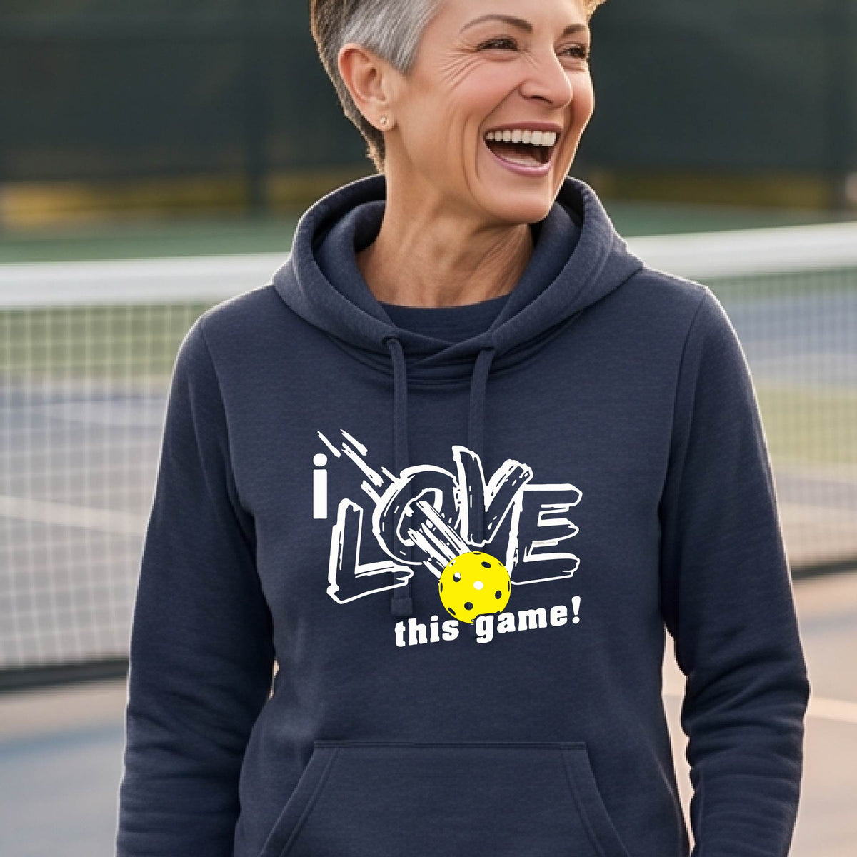 A woman wearing a navy blue hoodie with a graphic that reads 'i LOVE this game!' in white, with a yellow pickleball over the letter O. 