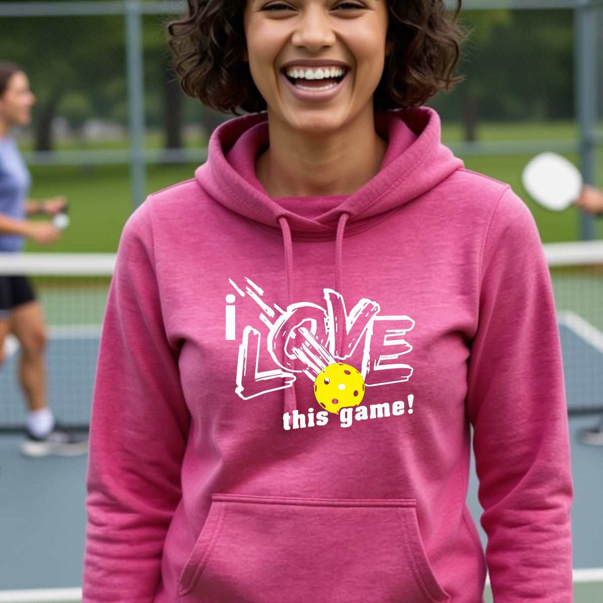 A woman wearing a pink hoodie with a graphic that reads 'i LOVE this game!' in white, with a yellow pickleball over the letter O. 