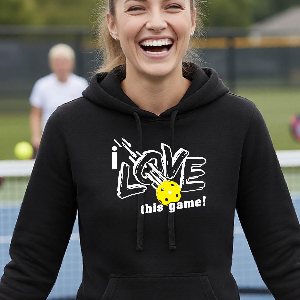 A woman wearing a black hoodie with a graphic that reads 'i LOVE this game!' in white, featuring a yellow pickleball in the center, on an outdoor pickleball court.
