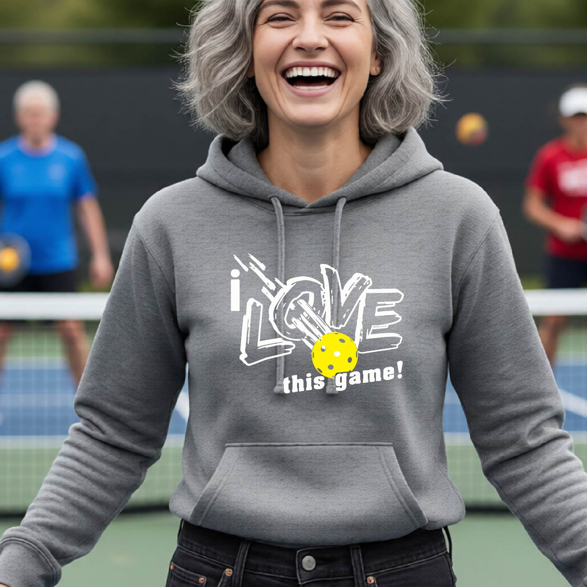 A woman wearing a gray hoodie with a graphic that reads 'i LOVE this game!' in white, featuring a yellow pickleball in the center, on an outdoor pickleball court.