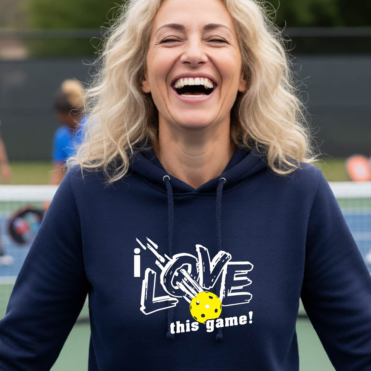 A woman wearing a navy blue hoodie with a graphic that reads 'i LOVE this game!' in white, featuring a yellow pickleball in the center, on an outdoor pickleball court.