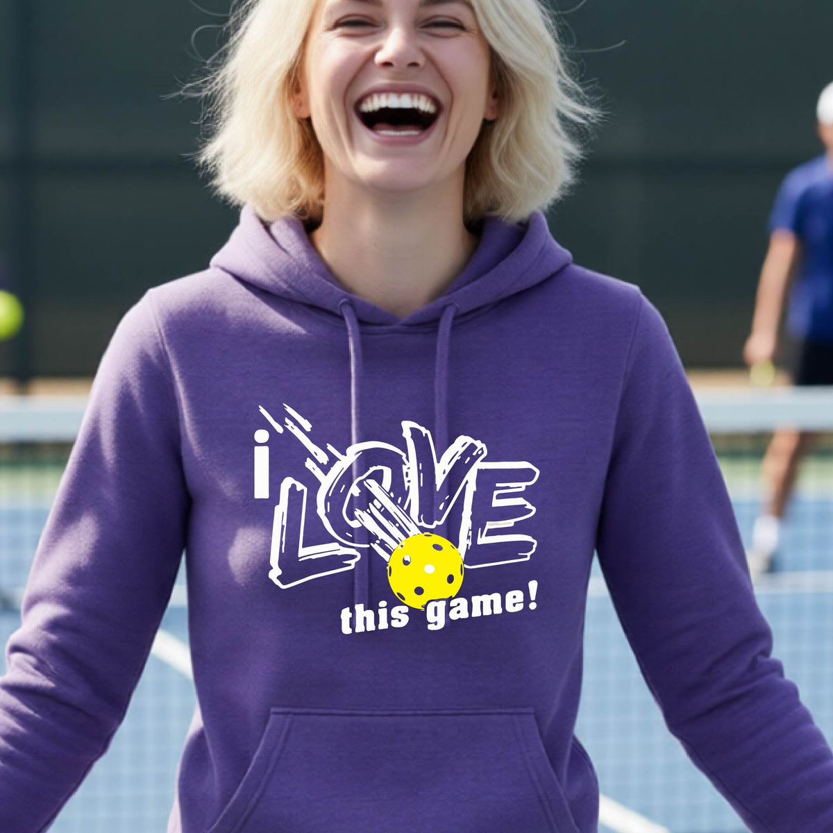 A woman wearing a purple  hoodie with a graphic that reads 'i LOVE this game!' in white, featuring a yellow pickleball in the center, on an outdoor pickleball court.