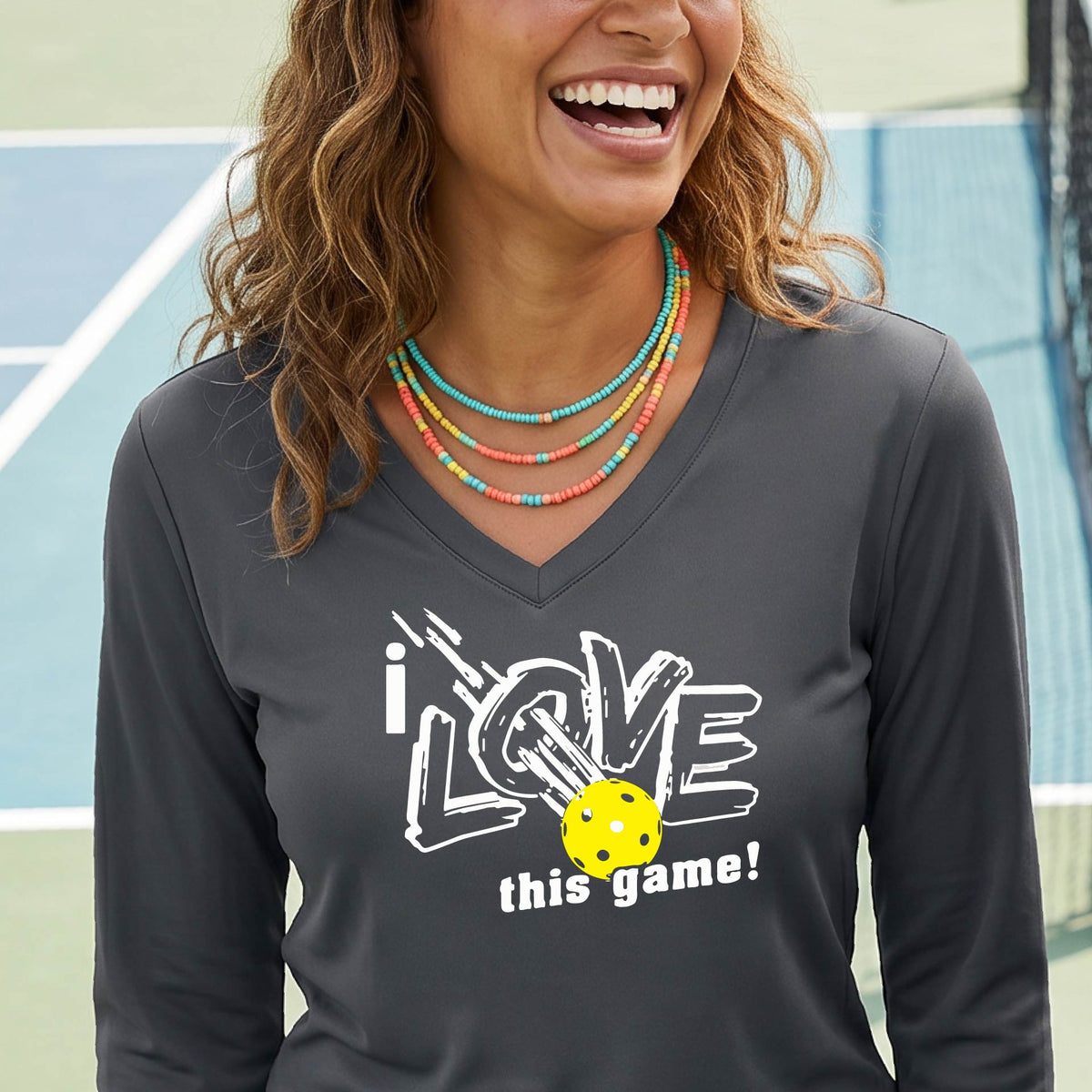 A woman on a pickleball court wearing a dark gray  long-sleeve V-neck athletic shirt with a white graphic that reads 'i LOVE this game!' and a yellow tennis ball illustration.