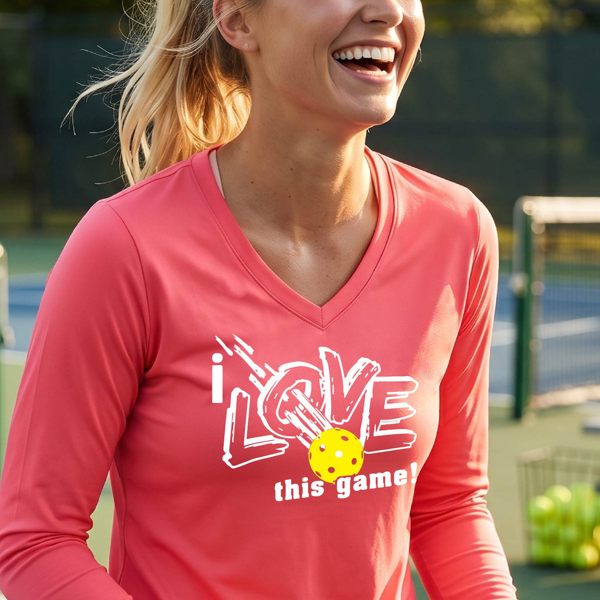 A woman on a pickleball court wearing a coral pink long-sleeve V-neck athletic shirt with a white graphic that reads 'i LOVE this game!' and a yellow tennis ball illustration.