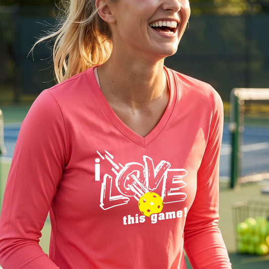 A woman on a pickleball court wearing a coral pink long-sleeve V-neck athletic shirt with a white graphic that reads 'i LOVE this game!' and a yellow tennis ball illustration.