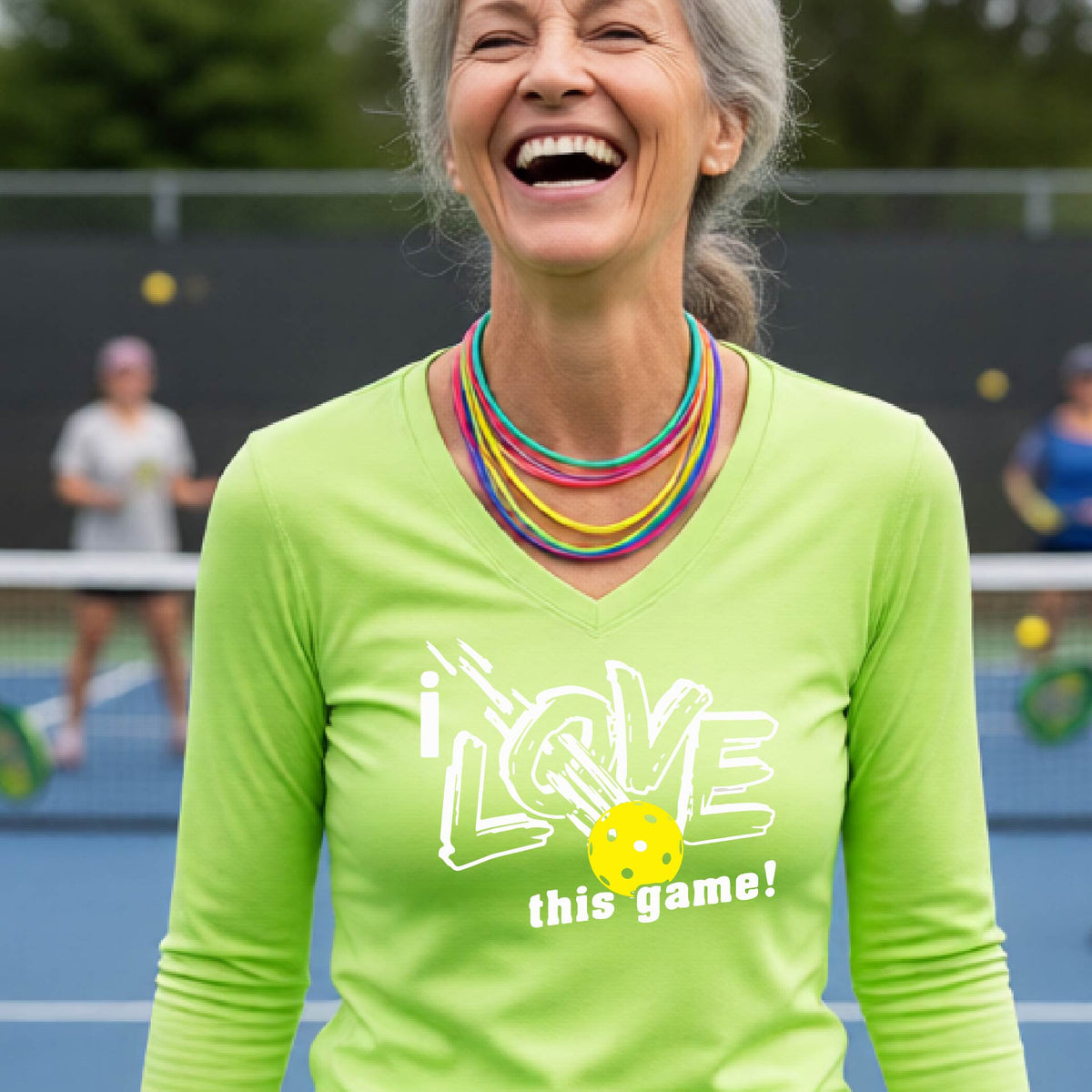 A woman on a pickleball court wearing a lime green long-sleeve V-neck athletic shirt with a white graphic that reads 'i LOVE this game!' and a yellow tennis ball illustration.